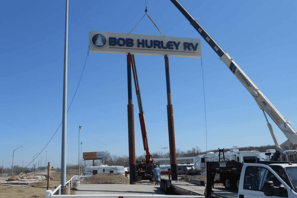 Sign reading 'Bob Hurley RV' being hoisted into place by a crane for a roadside RV sign installation.