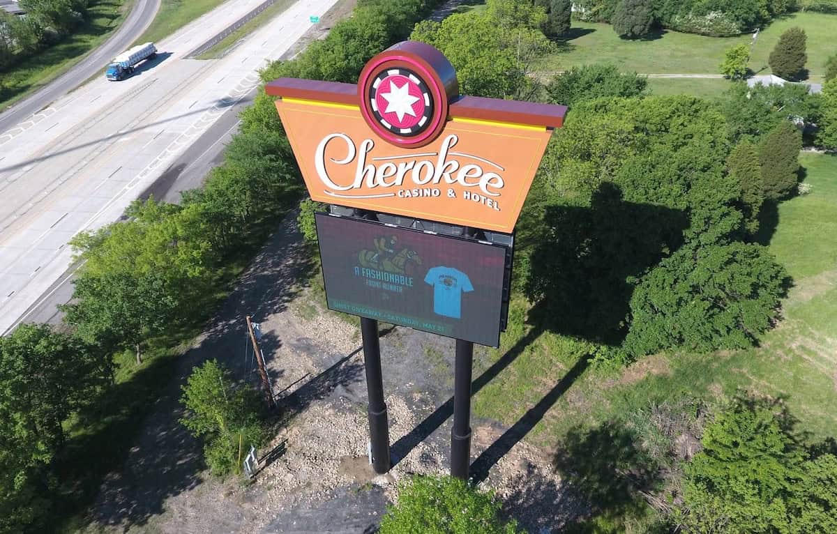 Aerial view of the Cherokee Casino & Hotel sign on tall posts beside a highway, with a digital billboard beneath and surrounding trees.